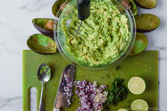 mashing avocado in a glass bowl with a potato masher and chopped onions, parsley, and cut lime for guacamole on a green cutting board below