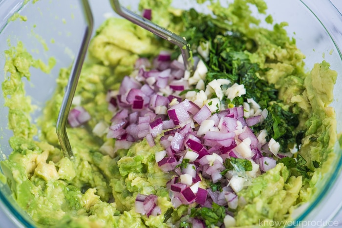 guacamole ingredients in a glass bowl ; avocado, garlic, cilantro, red onion, and lime juice