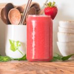 strawberry watermelon juice in a glass cup with stacked bowls to the back right and a cup with wooden spoons and reusable straws to the left and greenery around