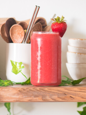 strawberry watermelon juice in a glass cup with stacked bowls to the back right and a cup with wooden spoons and reusable straws to the left and greenery around