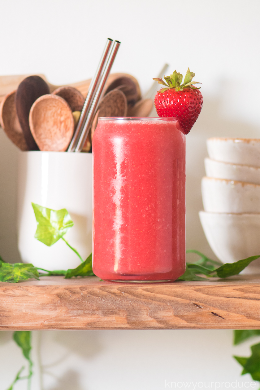 strawberry watermelon juice in a glass cup with stacked bowls to the back right and a cup with wooden spoons and reusable straws to the left and greenery around