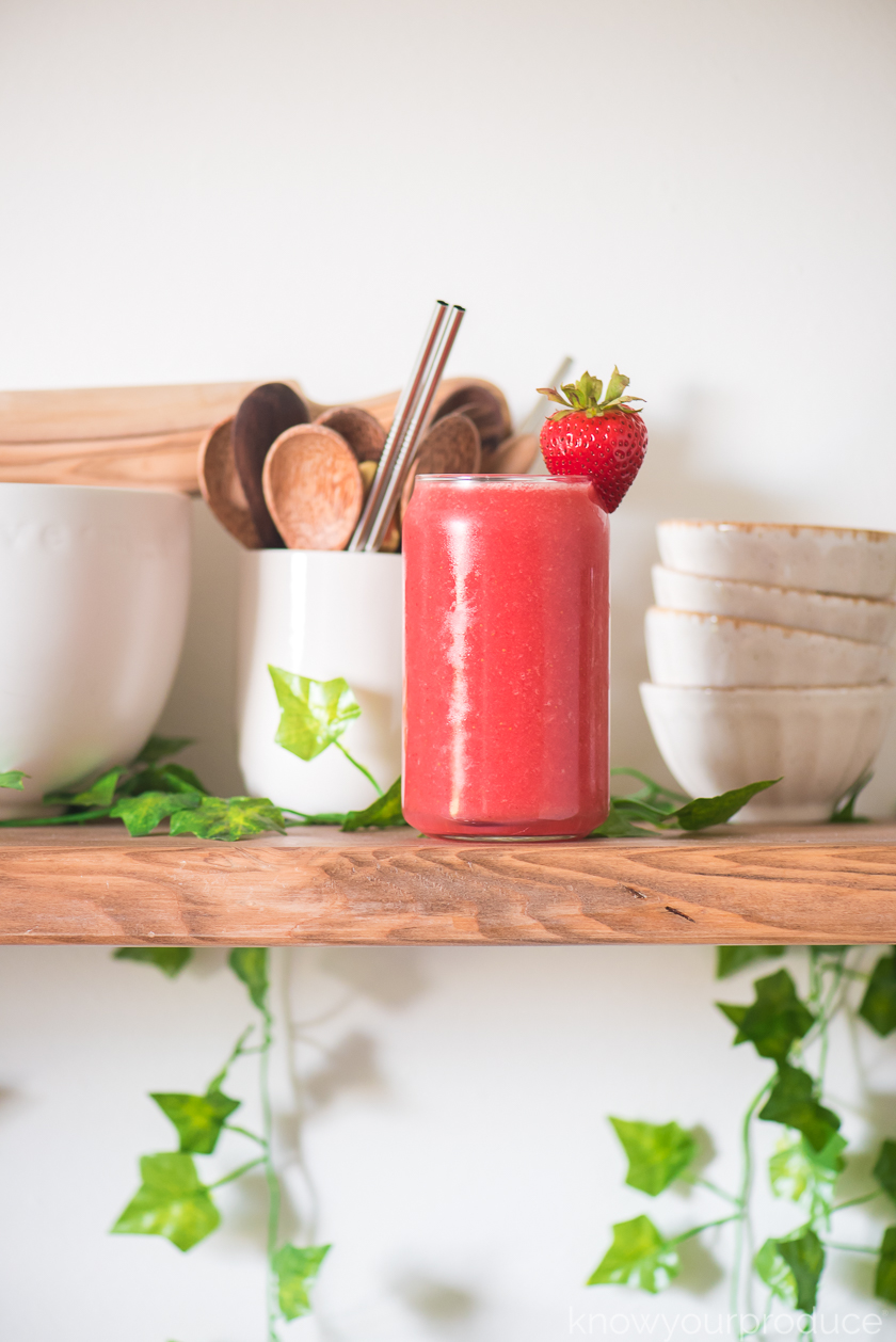 strawberry watermelon juice in a glass cup with stacked bowls to the back right and a cup with wooden spoons and reusable straws to the left and greenery around