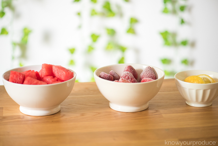 3 bowls on a table one with watermelon one with frozen strawberries and one with cut lemon wedges with greenery in the background