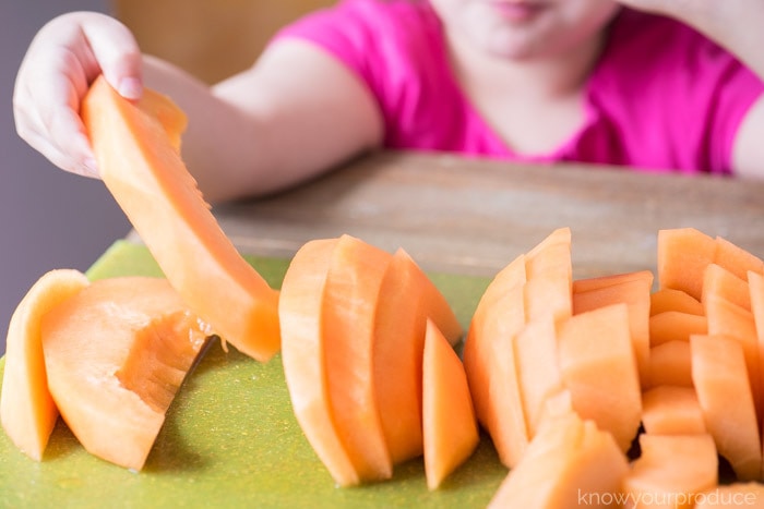 cut cantaloupe into wedges and chunks on a cutting board with child taking a slice