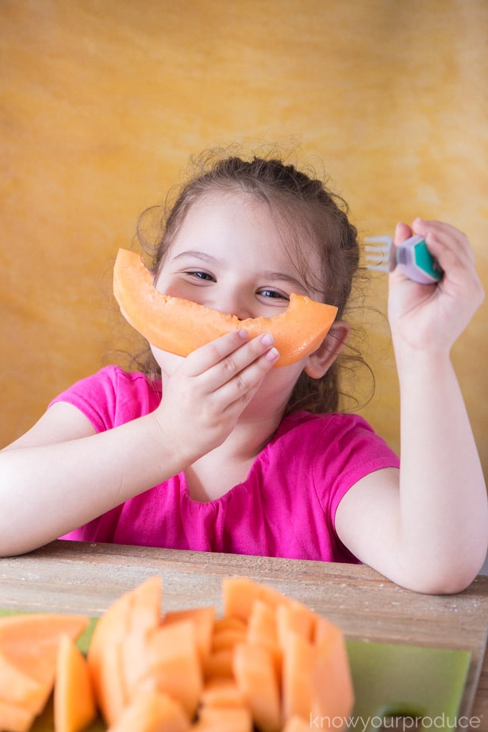 child holding cantaloupe wedge in hand acting as a smile by her face with fork in the other hand