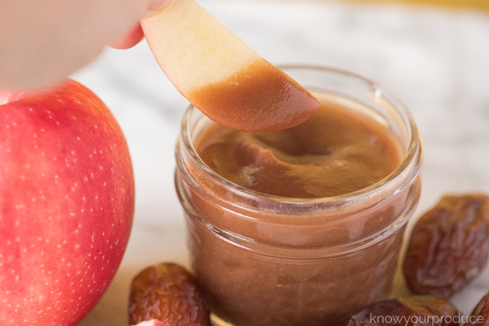 dipping apple into date butter spread in a mason jar next to apple with whole dates on marble