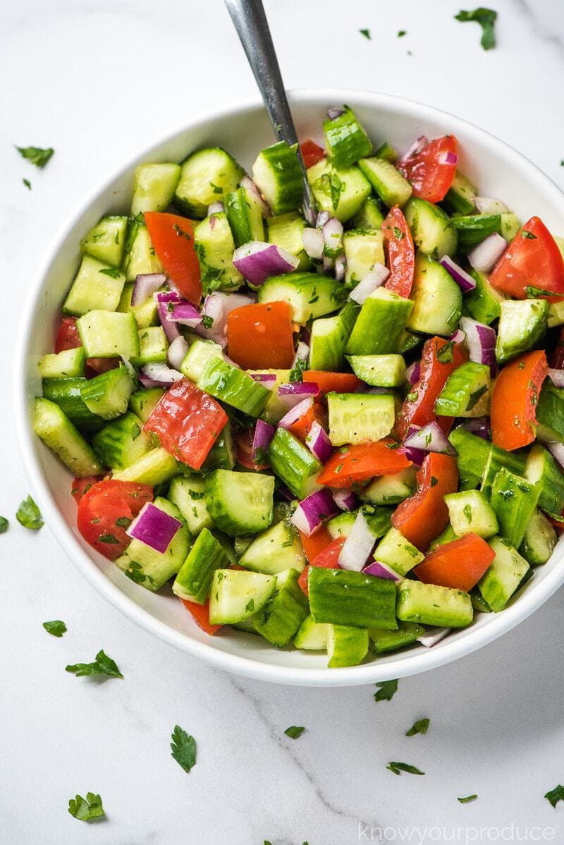 shirazi salad in a bowl with a spoon and fresh herbs scattered on a marble slab