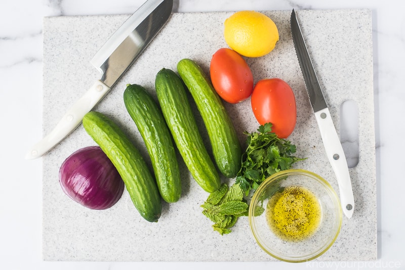 cucumbers, onions, tomatoes, lemon, oil, and herbs with knives on cutting board