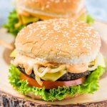 two vegan black bean burgers with mushrooms lettuce, tomato, burger sauce, caramelized onions, on a sesame seed bun on a round serving board with bark