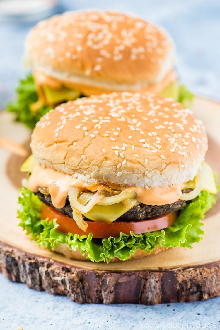 two vegan black bean burgers with mushrooms lettuce, tomato, burger sauce, caramelized onions, on a sesame seed bun on a round serving board with bark