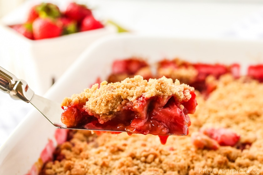 strawberry rhubarb crisp on a spatula with casserole dish and berries in background