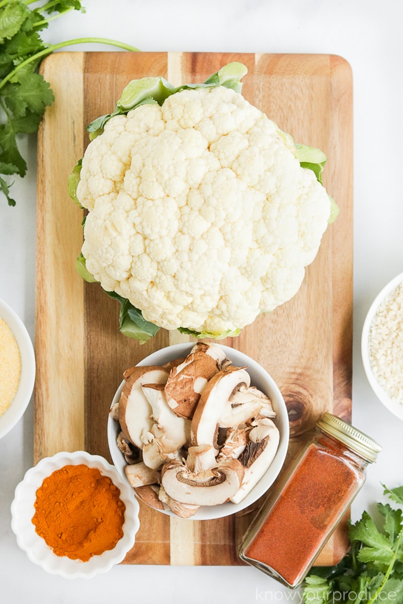 whole cauliflower with sliced mushrooms in a bowl and ingredients for cauliflower tacos on a cutting board