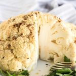whole roasted cauliflower with a piece cut out and parsley garnish on a white plate with fork to the side and gray and white striped cloth in background