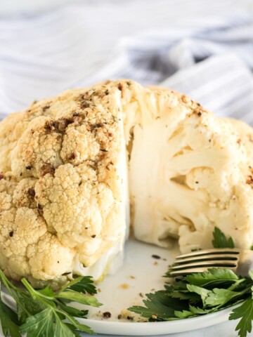 whole roasted cauliflower with a piece cut out and parsley garnish on a white plate with fork to the side and gray and white striped cloth in background