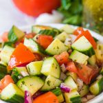 cucumber tomato salad with italian dressing in a white bowl with dressing in mason jar in background along with tomato parsley and garlic clove in background