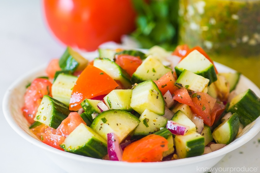 cucumber tomato salad in a low bowl with italian dressing in mason jar in background and tomato and parsley in back