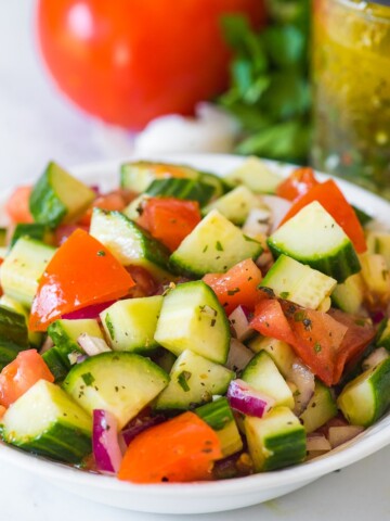 cucumber tomato salad with italian dressing in a white bowl with dressing in mason jar in background along with tomato parsley and garlic clove in background