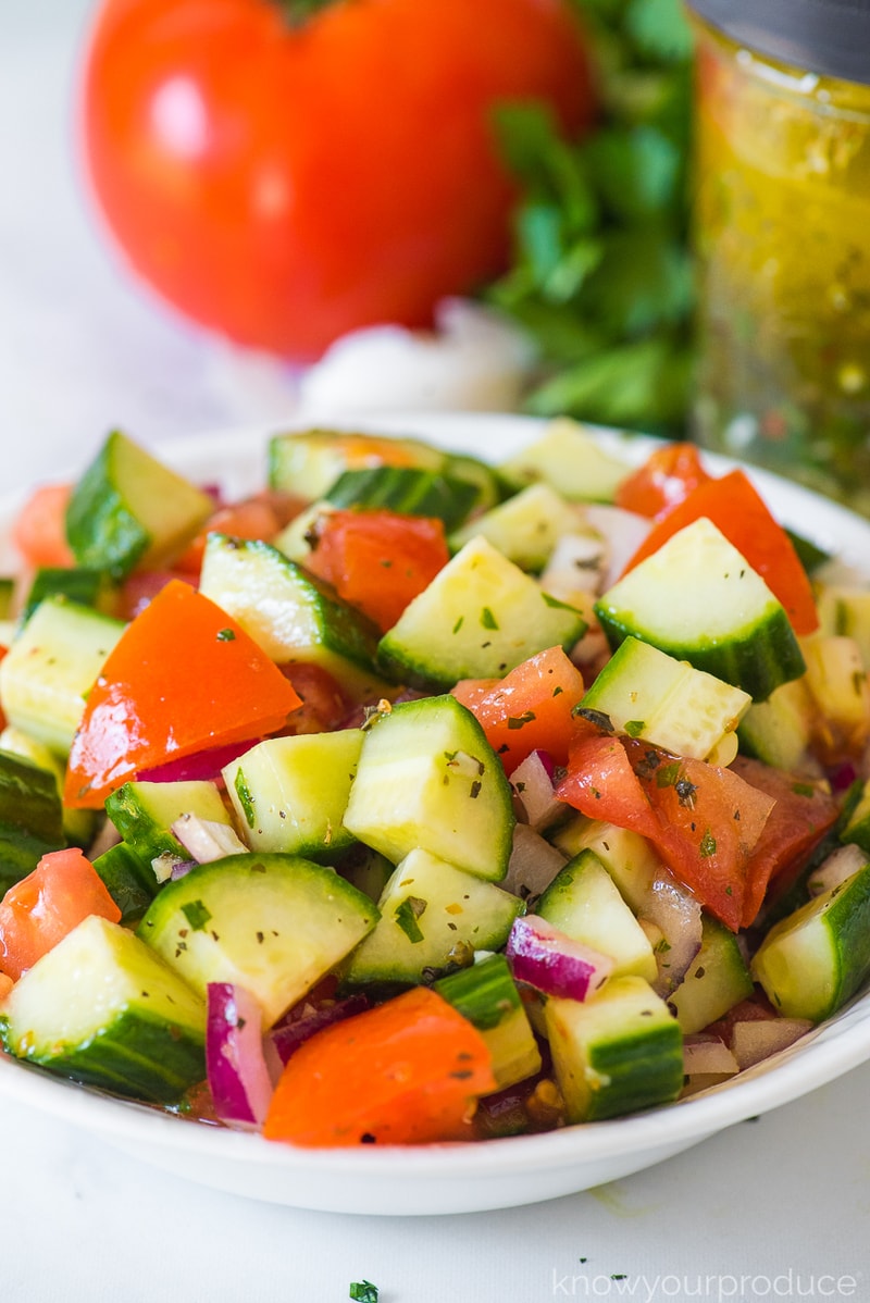 cucumber tomato salad with italian dressing in a white bowl with dressing in mason jar in background along with tomato parsley and garlic clove in background