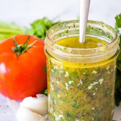 italian salad dressing in a mason jar with a spoon and tomato garlic clove and parsley behind it