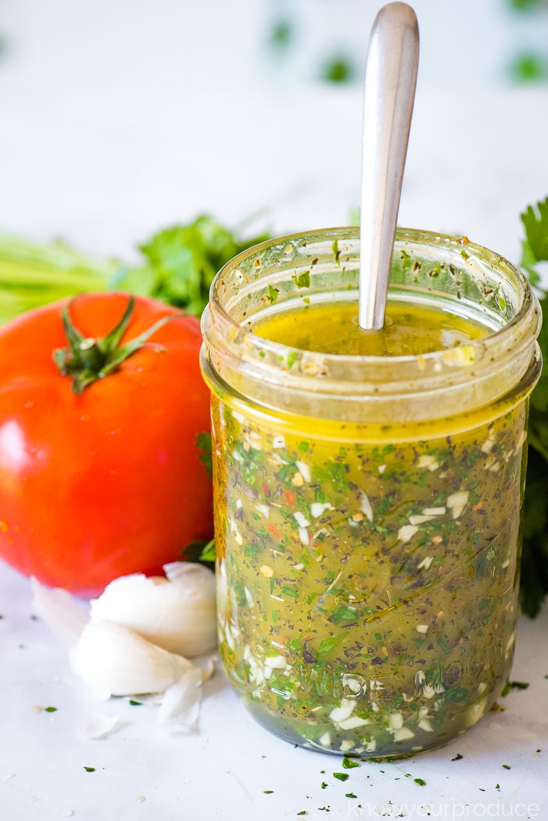 italian salad dressing in a mason jar with a spoon and tomato garlic clove and parsley behind it