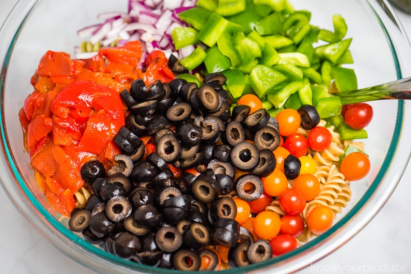 chopped vegetables olives whole petite cherry tomatoes over pasta in a large glass bowl with spoon to the right