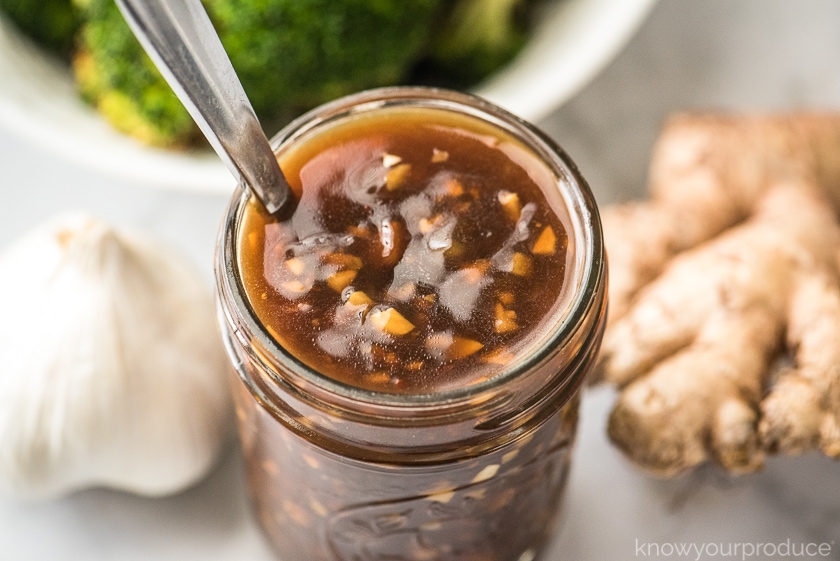 chinese garlic sauce in mason jar with garlic and ginger on table and broccoli in bowl in the back