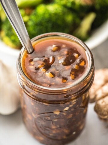 chinese garlic sauce in a mason jar with spoon and garlic to the left ginger to the right and broccoli in the back