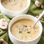 vegan cream of mushroom soup in small white bowls with mushrooms on green cloth with scattered mushrooms on it