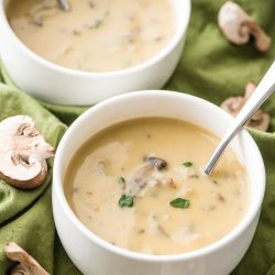 vegan cream of mushroom soup in small white bowls with mushrooms on green cloth with scattered mushrooms on it