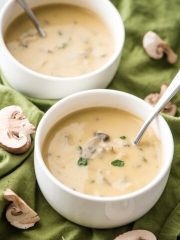 vegan cream of mushroom soup in small white bowls with mushrooms on green cloth with scattered mushrooms on it