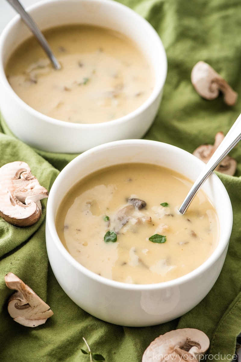 vegan cream of mushroom soup in small white bowls with mushrooms on green cloth with scattered mushrooms on it