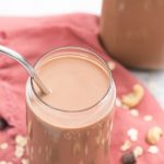 vegan chocolate oat milk with cashews in a glass cup with stainless steel straw on a pink cloth and cashews oats and chocolate scattered on napkin