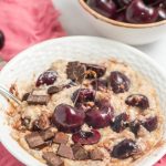 oatmeal with fresh cherries and chocolate chunks with spoon in a white low bowl on a pink napkin with cherries in a small ceramic bowl in the back right
