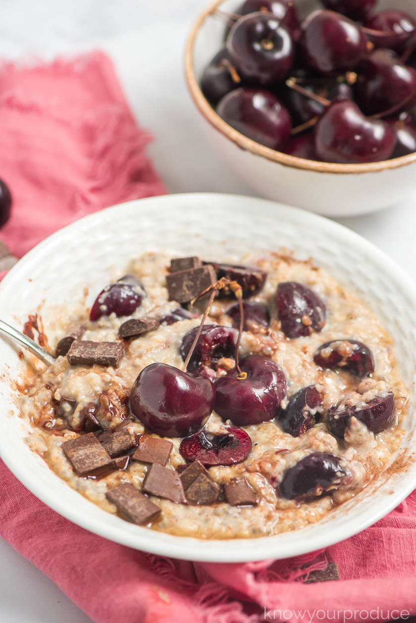 oatmeal with fresh cherries and chocolate chunks with spoon in a white low bowl on a pink napkin with cherries in a small ceramic bowl in the back right