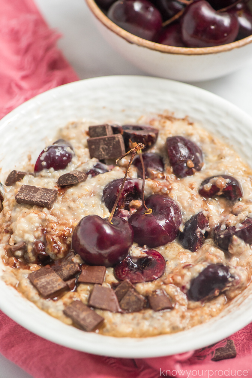 cherry oatmeal with fresh cherries and chocolate chunks with spoon in a white low bowl on a pink napkin with cherries in a small ceramic bowl in the back right