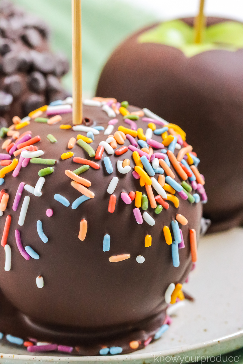 close up chocolate covered apple with sprinkles on a plate with two more apples and green napkin in the background