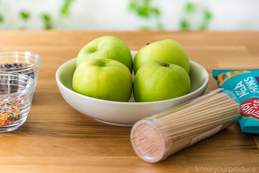 granny smith apples in a bowl with screwers and chocolate chips to the side and sprinkles and chocolate chips to the left in glass bowls on a table