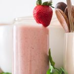 fresh strawberry peanut butter banana smoothie in a glass with strawberry on top on a wooden shelf with small bowls and cup with wooden spoons in background