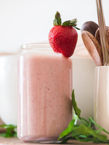 fresh strawberry peanut butter banana smoothie in a glass with strawberry on top on a wooden shelf with small bowls and cup with wooden spoons in background