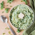 colcannon with tab of butter in a white low bowl with green napkin to the right and spoon to the left on tan backdrop with scattered kale and sliced green onions