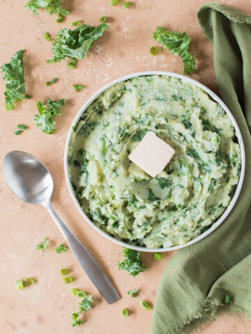 colcannon with tab of butter in a white low bowl with green napkin to the right and spoon to the left on tan backdrop with scattered kale and sliced green onions