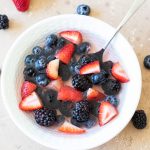 blueberries, strawberries chopped, blackberries, and coconut water in a white bowl with a spoon and berries scattered on a tan backdrop