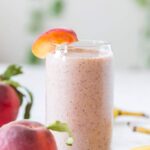 banana peach smoothie in a glass with a peach on the rim of the glass and 2 peaches on the left and banana on the right