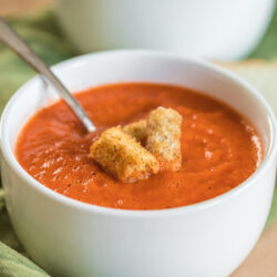 Roasted Red Pepper and Tomato Soup in a white small bowl with croutons and spoon on a green napkin