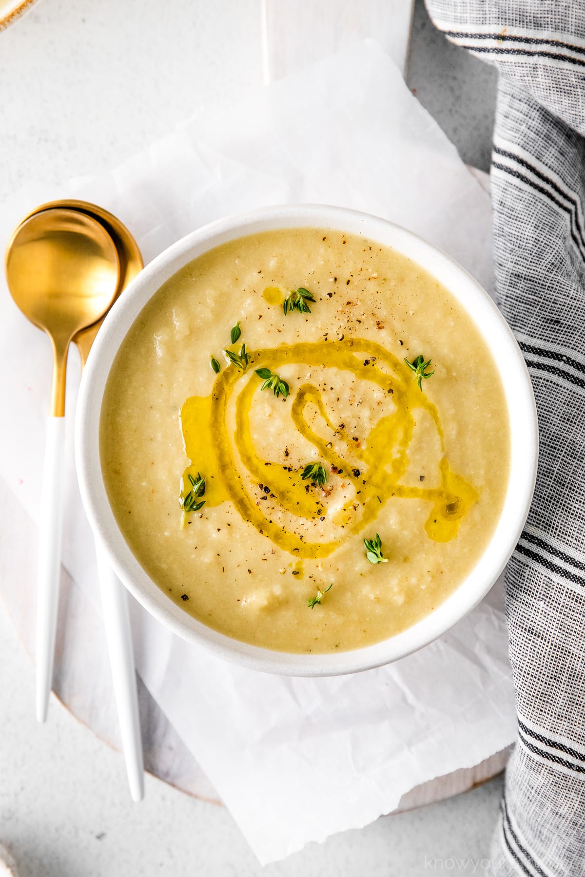 flat lay of cauliflower leek soup in a white bowl with olive oil drizzle and thyme garnish and gold plated spoons to the left gray striped napkin to the right