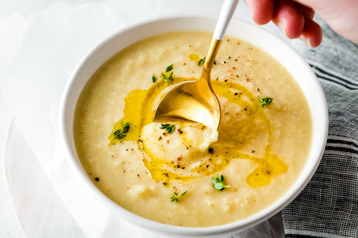 cauliflower leek soup in a white bowl with olive oil drizzle and thyme garnish with spoon being held by hand getting a bite