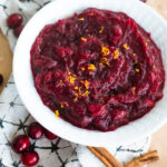 overhead fresh cranberry sauce in a white bowl with a napkin under it and cranberries and cinnamon sticks scattered