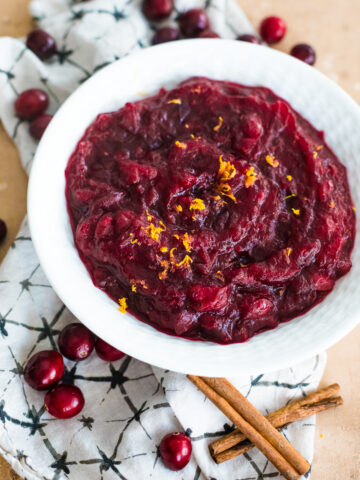 overhead fresh cranberry sauce in a white bowl with a napkin under it and cranberries and cinnamon sticks scattered