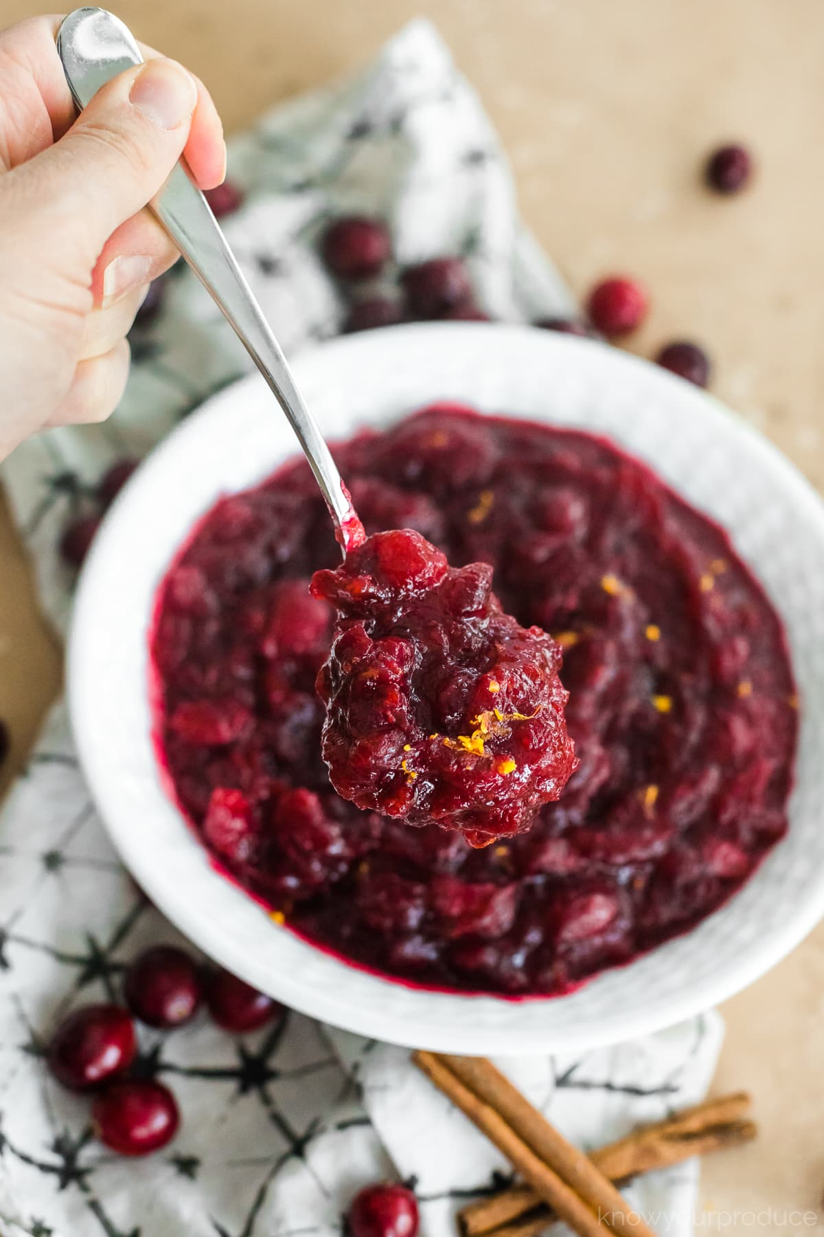 hand with spoon scooping fresh cranberry sauce in a white bowl with a napkin under it and cranberries and cinnamon sticks scattered