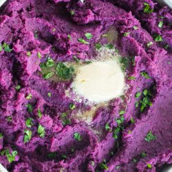 close up of mashed purple sweet potatoes with a pat of butter and parsley garnish in a white bowl with a spoon on the top